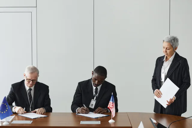 Executives signing international agreement with EU and US flags displayed on a wooden table. Aftaleindhold i strategiske partnerskaber