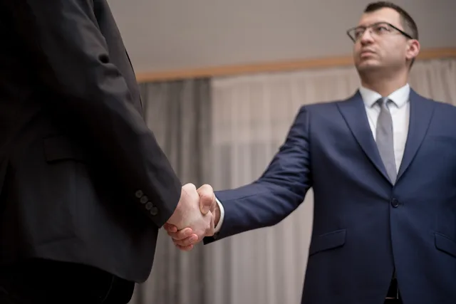 Two businessmen in formal attire shaking hands during a meeting. Partnerskaber mellem virksomheder: former, aftaler og fælles værdi